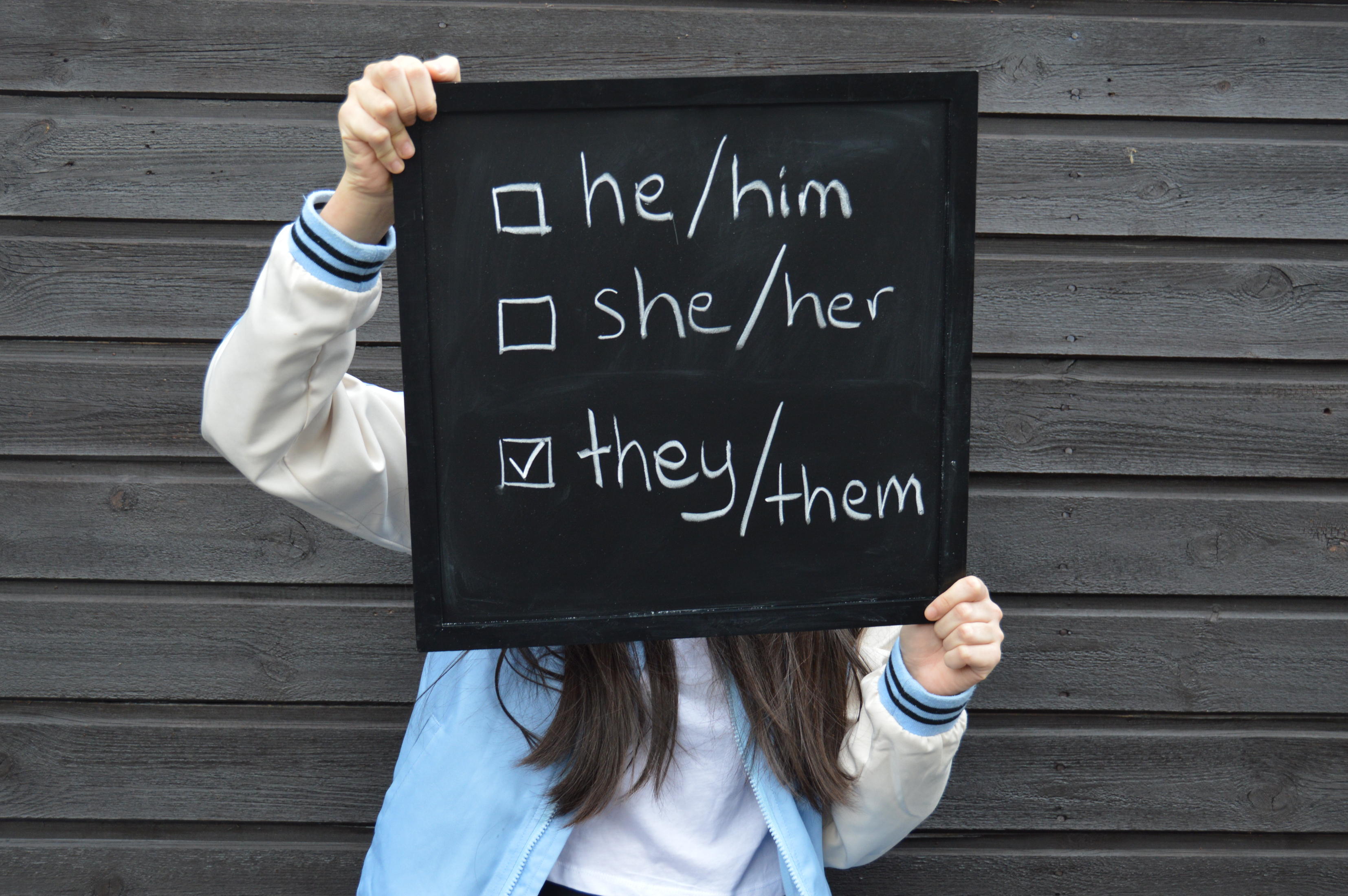 Young person in a blue and white jacket, holding a black sign with a short check-list that reads: he/him (box unchecked), she/her (box unchecked), they/them (box checked)