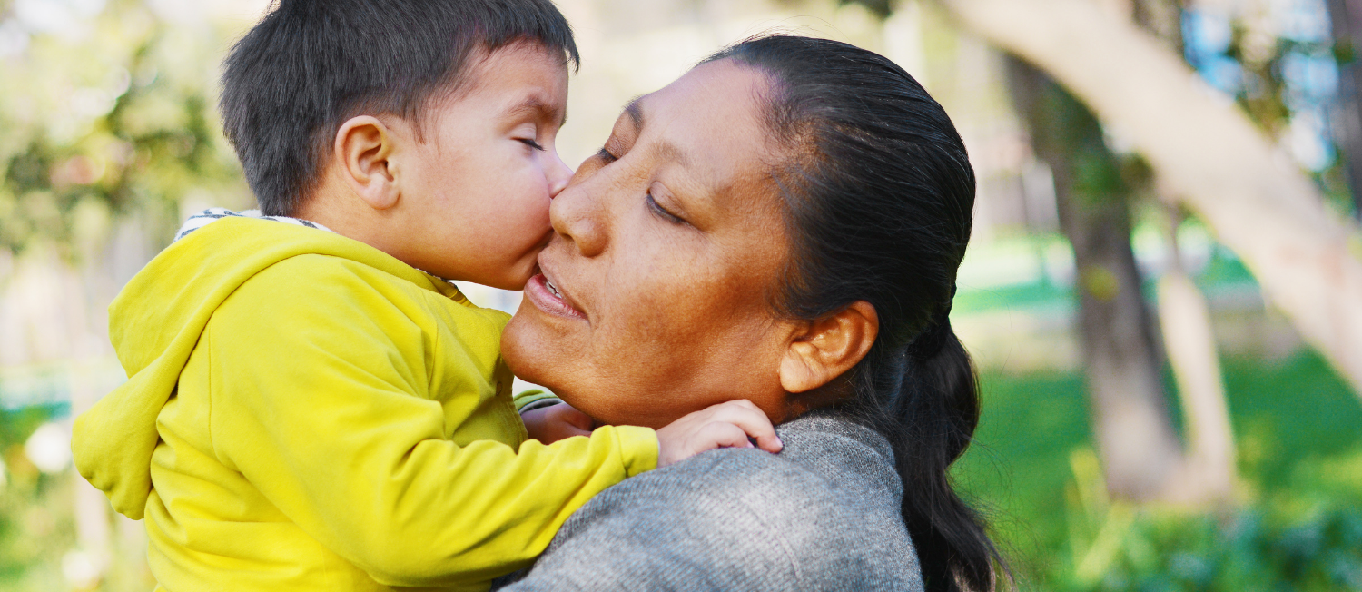 Photo of person with long dark hair being kissed on the cheek by a child in a bright yellow jacket