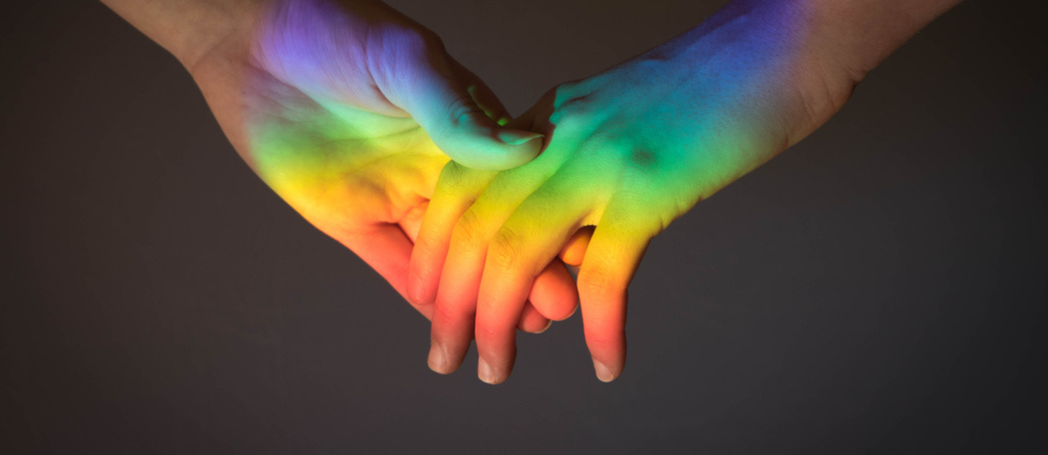 Close-up photo of two people holding hands, and rainbow light reflected on their hands.