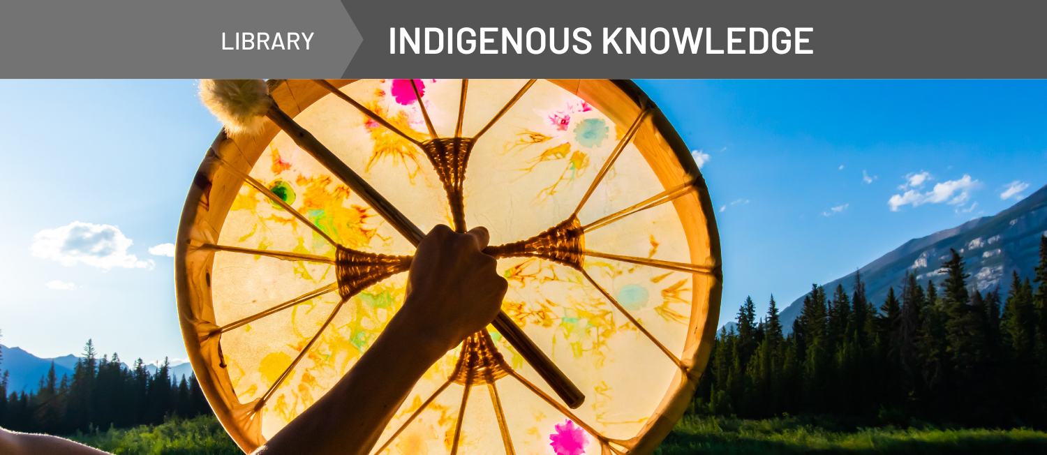 Photo of an Indigenous person holding a drum. Behind them is a scene of a lake and forested mountains.