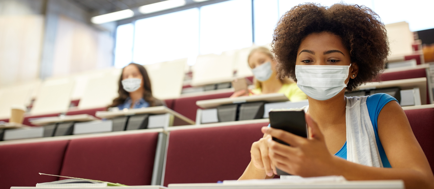Masked college student with dark coily hair, sitting in a large well-lit lecture hall