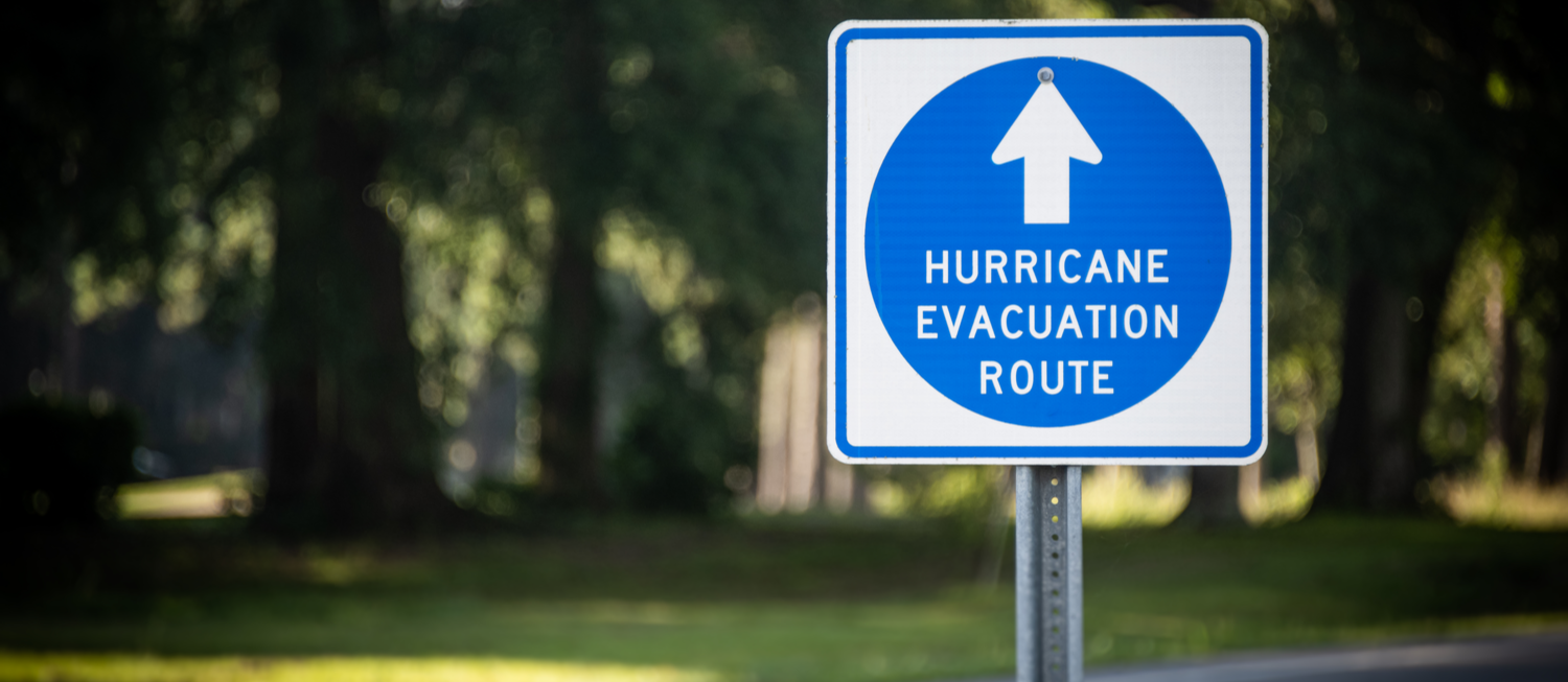 Photo of blue and white Hurricane Evacuation Route sign in front of trees and a road.